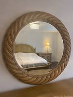 Front view of large round mirror hanging on a wall reflecting bedroom furniture and lamp.