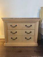 Front view of the wood veneer nightstand showing three drawers with decorative metal handles and carved details on the base and drawer frames.