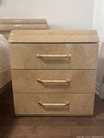 Front view of one of the matching veneer nightstands showing three drawers with brass handles and a glossy light beige finish.