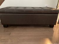 Front side view of the dark brown leather ottoman showing tufted button design and short wooden legs; placed on a wood floor.