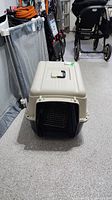 Front and top view of beige plastic pet carrier with black metal mesh front door. Handle visible on top.