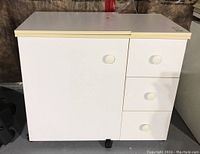 White laminate sewing machine cabinet closed, showing two leaves on top, one cabinet door and three drawers with round knobs, resting on black castors.