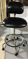 Full view of adjustable height black vinyl stool with backrest and chrome frame on caster wheels.