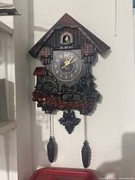 Full front view of the brown plastic cuckoo wall clock shaped like a chalet house with pinecone weights and a small bird above the clock face.