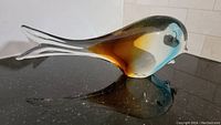 Photo of blown glass fish sculpture viewed side angled showing amber, blue, white, and clear colors and glossy finish on dark surface.