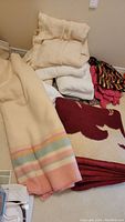 Stack of assorted blankets and throws piled on floor, showing various colors and textures including cream, red, white, and multi-colored.