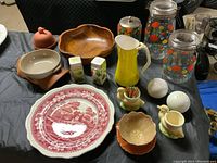 Overview of vintage kitchen items including wooden bowl, ceramic plate, enamel pitcher, canisters, salt and pepper shakers, and egg cups displayed on black tablecloth.