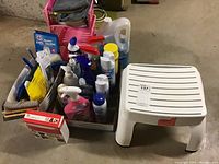 Photo of cleaning supplies including disinfectant sprays, cleaning liquids, soap bottles, cloths and scrubbers organized in containers on floor next to white plastic step stool.