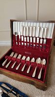 Opened wooden canteen showing multiple rows of knives, forks, and spoons with engraved handles in red velvet slots.