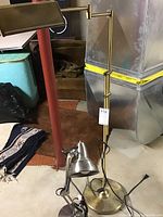 Brass floor lamp with adjustable arm and rectangular shade, standing on floor with electrical cord visible. Silver bendable desk lamp placed near the base of the floor lamp.