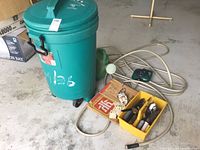 Green Rubbermaid garbage can with lid and wheels next to coiled garden hose and yellow container with hand tools, paper bags, and owl wind chime on garage floor