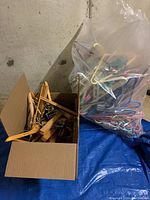Cardboard box holding wooden coat hangers and silver metal pant hangers with clips on a blue tarp.