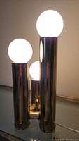 Three varying height brass-colored cylindrical pillars with white globe bulbs arranged atop, photographed from a frontal close angle to show lamp structure.
