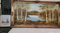Full view of both paintings together: large birch lakeside scene in an ornate frame and smaller jack pine winter scene