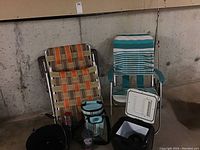Two folding web strap lounge chairs, one orange and brown and one turquoise and white, along with a small white electric cooler, soft cooler bag, thermos, and large black enamel pot arranged against a concrete wall.