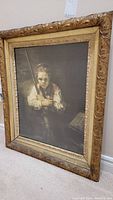 Framed print of a girl holding a broom, frame is ornate with floral carvings all around, overall frame size about 28x22 inches, positioned leaning against wall on floor.