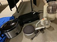 Photo of the small oval Crock-Pot slow cooker, electric griller, crepe maker, food processor and chopper on a basement floor showing the appliances together in storage.