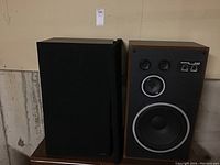 Pair of Sanyo AD 5075 speakers side by side on wooden stands in basement setting. One speaker with grille on, other grille off revealing drivers.