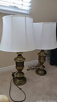 Two antiqued bronze color table lamps with flared white shades, positioned next to a window on carpeted floor. Both lamps have cords and visible finials but differ in harp structure under the shades.