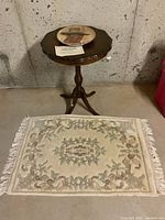 Photo showing the round wooden pedestal table with scalloped edges and brass claw feet, John Wayne plate on top, and wool mat on the floor beneath.