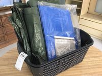 Black plastic tote bin holding two tarps, the green tarp is folded and loose, the blue tarp is new in packaging.
