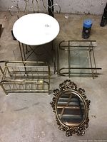Overview of all items on concrete floor: white seat stool, metal magazine rack, metal wall shelf with glass shelves, ornate oval mirror