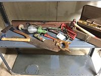 Full view of vintage tools laid out on metal shelf including saw, axe, hand drill, pipe wrenches, toolbox and tool roll