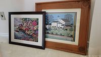 View of the framed needlepoint art of cottage by waterfront in dark frame alongside the larger double matted Charles Wysocki print titled 'Amish Meeting' in pine folk style frame with heart carvings.