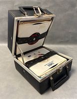 Black carrying case open displaying vinyl records secured by elastic strap inside.