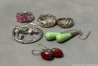 Photo showing the full lot of 6 jewelry pieces on grey leather surface including rings, brooch, and earrings.