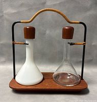 Front view of cruet set showing opaque white and clear glass decanters with wooden stoppers on teak tray with metal and rattan handle.