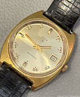 Front view of the Elgin vintage wristwatch showing gold-tone case, silver dial, date window, and black leather strap.