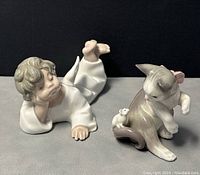 Two Lladro porcelain figurines: cat with mouse and lying angel, on gray surface with black backdrop