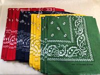 Photo showing four stacks of folded bandannas in red, navy blue, yellow, and green with white paisley patterns, made of 100% cotton.