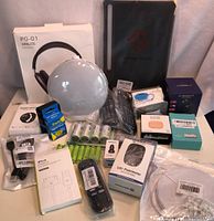Photo of multiple electronic items including white globe light, black wireless headphones, battery packs, pedometer boxes, and other electronic accessories packed on a surface.