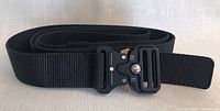 Black nylon men's belt coiled with metal quick-release buckle on display on a light textured surface.