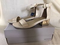 Side angle of a single new Kenneth Cole women's sandal on its box showing light beige color and gold block heel.