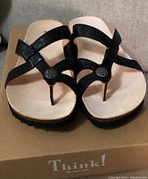 Front view of the black leather sandals showing toe loop and metallic button detail on straps placed on cardboard box with Think! logo.