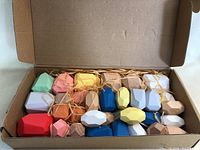 Open box showing 24 wooden stone-shaped building blocks in multiple colors including red, pink, yellow, blue, white, natural wood, and gray, resting on packing material inside a cardboard box.