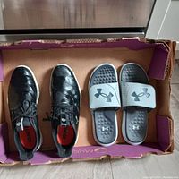 Photo of both pairs of footwear in a cardboard shoe box: black men's IGNITE leather shoes and gray Under Armour slippers, both size 10.5.