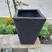 Top and angled view of the black wicker style plastic planter showing the square tapered shape and woven texture outdoors.