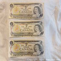 Photo showing three Canadian one dollar paper bills laid out flat, displaying serial numbers and Queen Elizabeth II portrait.