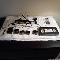 Electric hair clipper with power cord and five attachment combs arranged on white towel, including VHS tape and manual