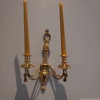 Photo showing a golden metal two-arm candelabra mounted on a wall, holding two gold taper candles. Visible decorative tassels and detailed metallic finish.