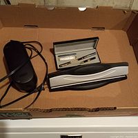 Photo showing the electric pencil sharpener with attached power cord, metal hole puncher, and boxed pen set with two pens.