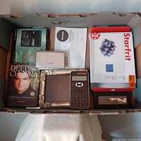 Box containing various electronics and kitchen items including Starfrit nutritional kitchen scale, calculators, electronic dictionary, Deepak Chopra DVD, picture frame, and bottle opener.