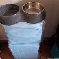 Two stacks of extra large dog pee pads on floor with a heavy plastic dual dog bowl holder on top holding one stainless steel bowl.