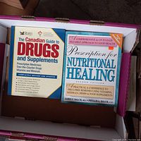 Two paperback books inside a cardboard box, titled 'The Canadian Guide to Drugs and Supplements' and 'Prescription for Nutritional Healing, Second Edition'. Both visible on top of each other.