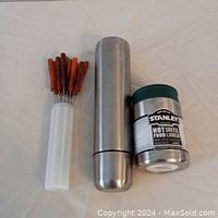 Photo showing two thermoses: one tall stainless steel thermos and one shorter Stanley brand insulated food thermos, along with a container holding 12 long metal skewers with wooden handles.