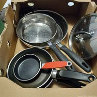Overview of six kitchen frying pans inside a cardboard box including two stainless steel with long black handles and three non-stick pans. Glass lid is visible.